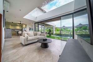 A modern living room with a sofa, coffee table, and a view of a swimming pool and greenery. - Luxe Jungle 4BR in the Heart of Ubud w Pool, Patio (Gianyar Regency)