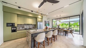 A modern kitchen with a central island, bar stools, and a dining area with a view of greenery. - Luxe Jungle 4BR in the Heart of Ubud w Pool, Patio (Gianyar Regency)