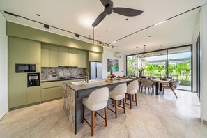 A modern kitchen with a central island, bar stools, and a dining area with a view of greenery. - Luxe Jungle 4BR in the Heart of Ubud w Pool, Patio (Gianyar Regency)