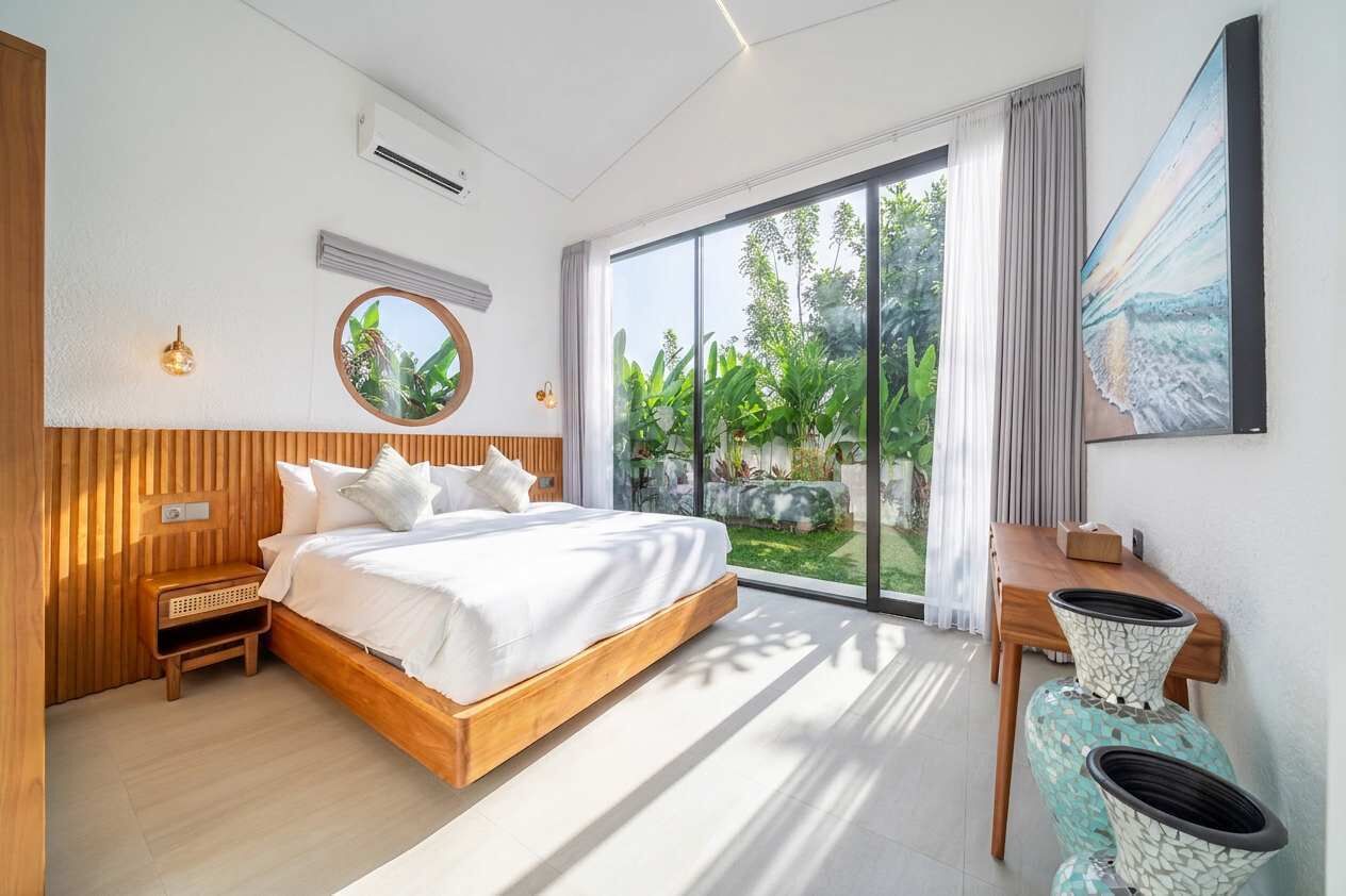 Une chambre moderne avec un grand lit, une tête de lit en bois, une table de chevet et une vue sur la verdure grâce à une porte-fenêtre coulissante.