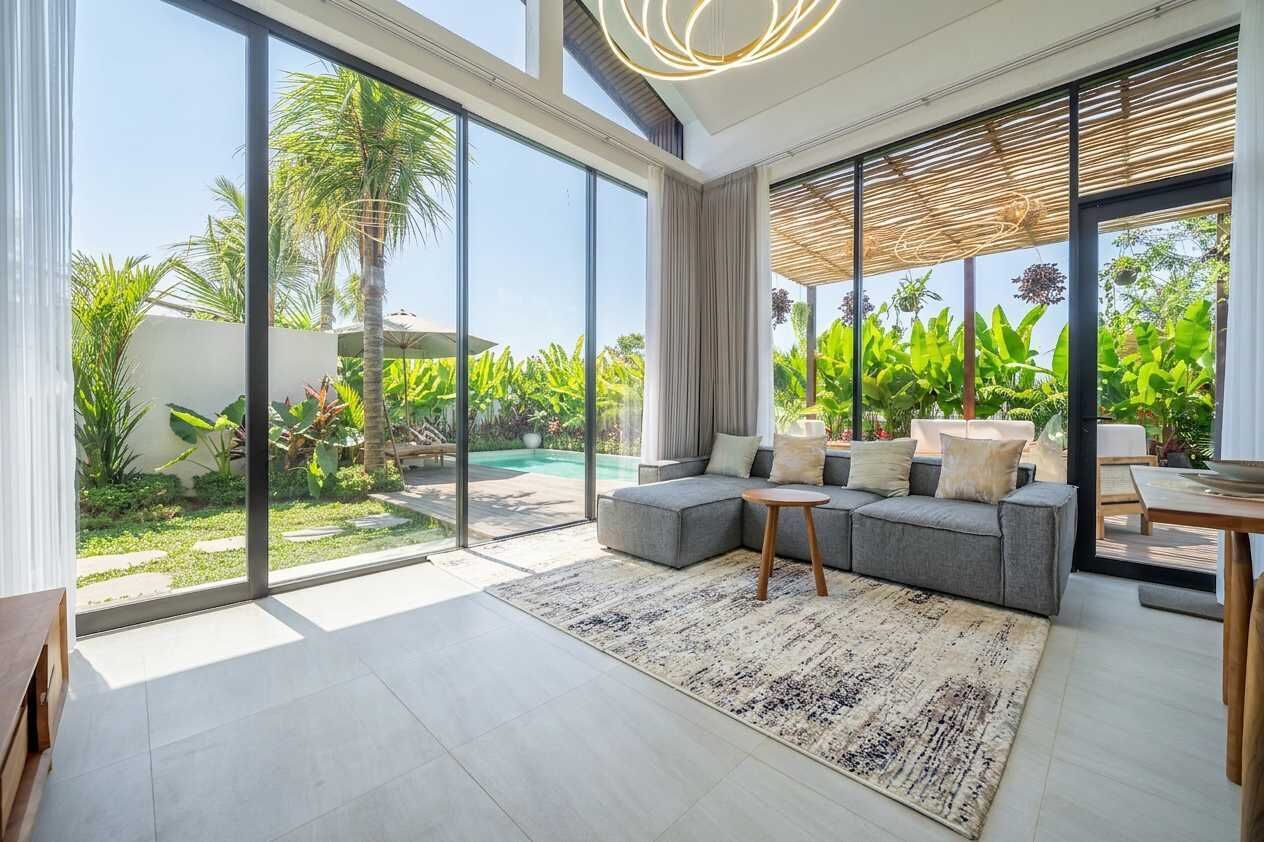 Un salon moderne avec un canapé d’angle gris, une petite table basse en bois et une grande fenêtre donnant sur une piscine et une végétation luxuriante.