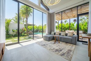 A modern living room with a grey sectional sofa, a small wooden coffee table, and a large window overlooking a pool and lush greenery. - Tropical 3BR w Sauna by Pandawa Beach • Pool+Patio (South Kuta)