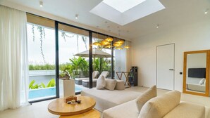 A modern living room with a sofa, a round wooden table, and a large window with a view of greenery and a pool. - Serene 2BR Villa in Ungasan- Close to Beach + Pool (Badung Regency)