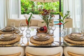 A dining table set with plates, bowls, and glasses, surrounded by chairs, with a view of greenery outside. - Designer 2BR in Ungasan w Pool & Beach Access (Badung Regency)