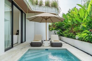 Un espace aménagé au bord de la piscine, avec deux chaises longues et un grand parasol, entouré d’une végétation luxuriante et bordé par la façade d’un bâtiment moderne.