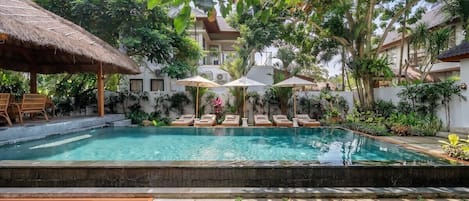 A swimming pool with lounge chairs and umbrellas, surrounded by lush greenery and a building in the background.