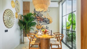 A dining area with a round wooden table, chairs, a large potted plant, and wall art. - Designer 2BR Villa in Serene Ubud w Pool & Garden (Gianyar Regency)