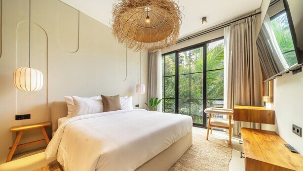A modern bedroom with a large bed, a wooden bedside table, a television, and a view of greenery through the window. - Designer 2BR Villa in Serene Ubud w Pool & Garden (Gianyar Regency)