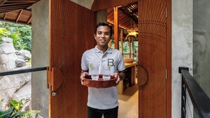 A person holding a tray with bottles, standing in a doorway with a wooden door and a view of greenery outside. - Jungle Villa, Brand New, Unique Art Design, Pool (Gianyar Regency)