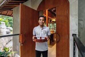 A person holding a tray with bottles, standing in a doorway with a wooden door and a view of greenery outside. - Jungle Villa, Brand New, Unique Art Design, Pool (Gianyar Regency)