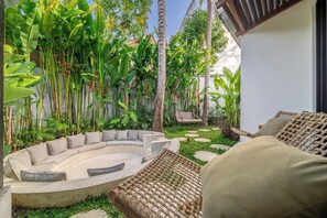 A circular outdoor seating area with cushions and a wicker chair, surrounded by lush tropical plants and a white wall. - Huge Tropical 3BR in Serene Ubud w Pool & Lounge (Ubud)