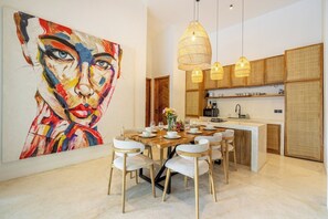A modern dining area with a large abstract painting, a wooden dining table, white chairs, and a kitchen in the background. - Huge Tropical 3BR in Serene Ubud w Pool & Lounge (Ubud)