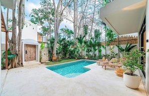 Ein moderner Poolbereich im Freien mit einem geometrisch gestalteten Pool, umgeben von üppiger Vegetation und Bäumen.