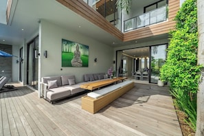 A modern outdoor living area with a sofa, bench, and dining table, surrounded by greenery and a large window. - Chic 4BR Villa w/ Private Massage Room & Huge Pool (Badung Regency)