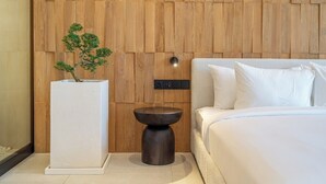 A bedroom with a wooden panel wall, a white bedside table with a potted plant, a round black side table, and a bed with white bedding. - New Tropical 3BR Villa+Private Pool+Best Location (South Kuta)