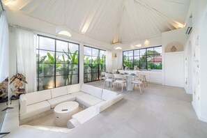 A spacious living area with a white sofa, a round coffee table, and a dining area with a white table and chairs. Large windows provide views of greenery outside. - Luxe 3BR Villa • Beach Nearby • Pool • Prime Spot (Badung Regency)