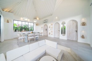A spacious living area with a high ceiling, a dining table with chairs, a white sofa, and a large window offering a view of greenery. - Luxe 3BR Villa • Beach Nearby • Pool • Prime Spot (Badung Regency)