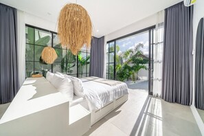 A modern bedroom with a large bed, a hanging light fixture, and a view of greenery outside. - Luxe 3BR Villa • Beach Nearby • Pool • Prime Spot (Badung Regency)