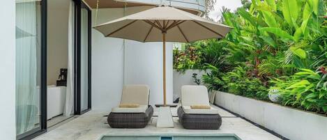 Uma área junto à piscina com duas espreguiçadeiras e um grande guarda-sol, cercada por vegetação exuberante e fachada de um edifício moderno.