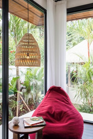 A room with a large window, a wicker pendant light, a red bean bag, and a small table with a book and decorative item. - Luxe 2BR Villa in Ubud w Garden Bath, Patio & Pool (Gianyar Regency)