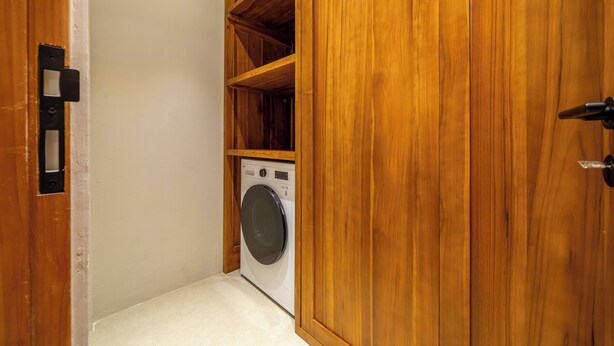 A laundry room with a washing machine, wooden shelving, and a door with a handle. - Cozy 3BR Villa in Lush Ubud w Pool, Patio & Garden (Gianyar Regency)