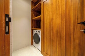 A laundry room with a washing machine, wooden shelving, and a door with a handle. - Cozy 3BR Villa in Lush Ubud w Pool, Patio & Garden (Gianyar Regency)
