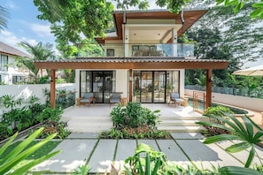 A modern two-story house with a covered patio, outdoor seating, and lush greenery.