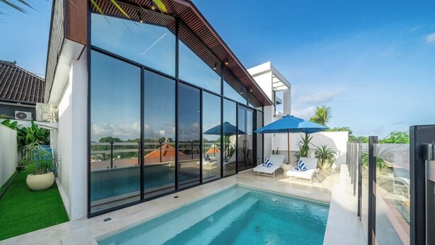 Un área de piscina exterior moderna con paredes de vidrio, sillones de descanso y sombrillas.