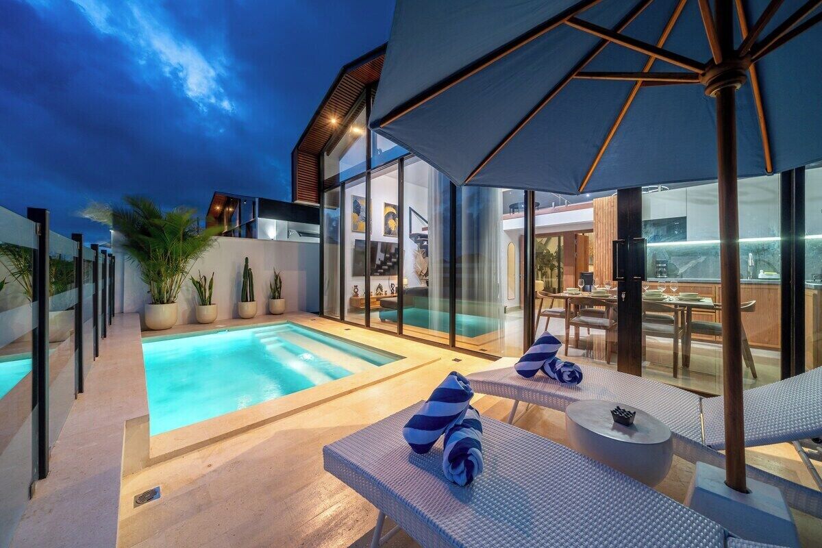 A modern outdoor pool area with lounge chairs and umbrellas, surrounded by glass walls and potted plants.