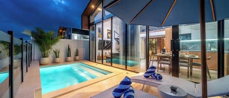 Un área de piscina moderna al aire libre, con sillones reclinables y sombrillas, rodeada de paredes de cristal y plantas en macetas.