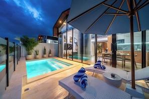 A modern outdoor pool area with lounge chairs and umbrellas, surrounded by glass walls and potted plants.