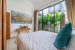 A bedroom with a large bed, a painting on the wall, a window with plants, and a wooden bedside table.