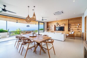 A modern living space with a dining area, kitchen, and seating area, featuring large windows and a view of the outdoors. - Luxe 4BR Villa w Rooftop, Cinema, Ocean View (Badung Regency)
