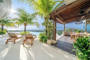 A covered outdoor seating area with lounge chairs and a table, surrounded by potted plants and palm trees, overlooking a scenic view. - Luxe 4BR Villa w Rooftop, Cinema, Ocean View (Badung Regency)