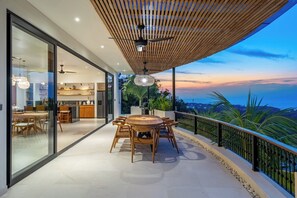 A modern outdoor dining area with a wooden table and chairs, a ceiling fan, and a view of the sunset over the ocean. - Luxe 4BR Villa w Rooftop, Cinema, Ocean View (Badung Regency)