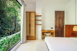 A bedroom with a bed, a desk, and a wardrobe. There is a window with a view of greenery outside. - Cozy 2BR Flat in Ubud's Centre w Pool & Kitchen (Ubud)