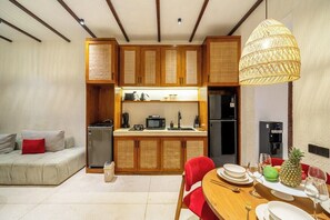 A compact kitchen with wooden cabinets, a refrigerator, and a dining area with a table set for two. - Cozy 2BR Flat in Ubud's Centre w Pool & Kitchen (Ubud)