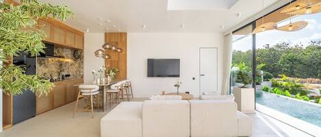 Uma sala de estar moderna com um amplo sofá em formato de L, um espaço de jantar com banquetas, uma cozinha com armários de madeira e uma grande janela com vista para uma piscina.