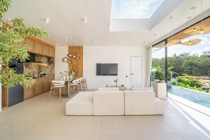 Ein modernes Wohnzimmer mit einem großen Ecksofa, einem Essbereich mit Barhocker, einer Küche mit Holzschränken und einem großen Fenster mit Blick auf einen Pool.