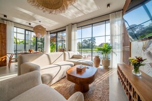 Ein modernes Wohnzimmer mit großen Fenstern, einer Sofagarnitur, einem Couchtisch und einer Holzkommode.
