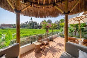 A covered outdoor seating area with a view of a rice field, a bamboo structure, and a potted plant with pink flowers. - Tropical 2BR in Ubud's Ricefields w Pool & Garden (Tegallalang)