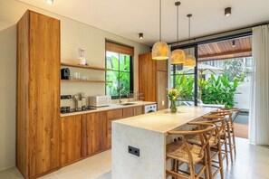 A modern kitchen with wooden cabinets, a central island, and a view of greenery outside. - Serene 2BR Retreat w Spacious Pool & Garden • Ubud (Gianyar Regency)