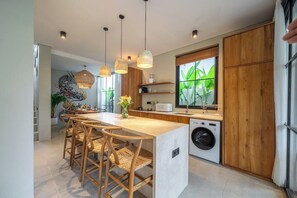 A modern kitchen with a central island, wooden chairs, a washing machine, and a large window with greenery. - Serene 2BR Retreat w Spacious Pool & Garden • Ubud (Gianyar Regency)