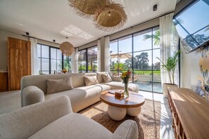 A modern living room with a sofa, coffee table, and large windows offering a view of the outdoors. - Tranquil 2BR Villa in Ubud • Pool • Patio • Garden (Gianyar Regency)