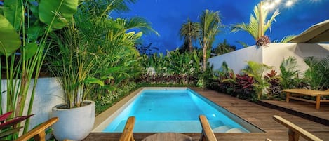 Una piscina rodeada de plantas tropicales, sillas de madera y una planta en maceta.