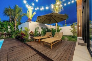 Une terrasse en bois avec deux fauteuils de jardin en osier et un grand parasol, entourée d’une végétation tropicale luxuriante et d’une piscine.