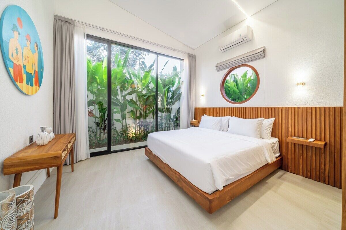 Ein Hotelzimmer mit einem Bett, einem Holz-Kopfteil, einem runden Spiegel, einem kleinen Tisch und einem großen Fenster mit Blick auf Grünflächen.