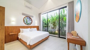 A bedroom with a bed, a wooden headboard, a bedside table, a wall-mounted air conditioner, a window with a view of greenery, and a circular wall decoration.