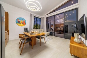 A modern living room with a dining area, a large TV, and a glass door leading to a balcony. - New Jungle 3BR Villa+Beach Access+Huge Pool 10A (South Kuta)
