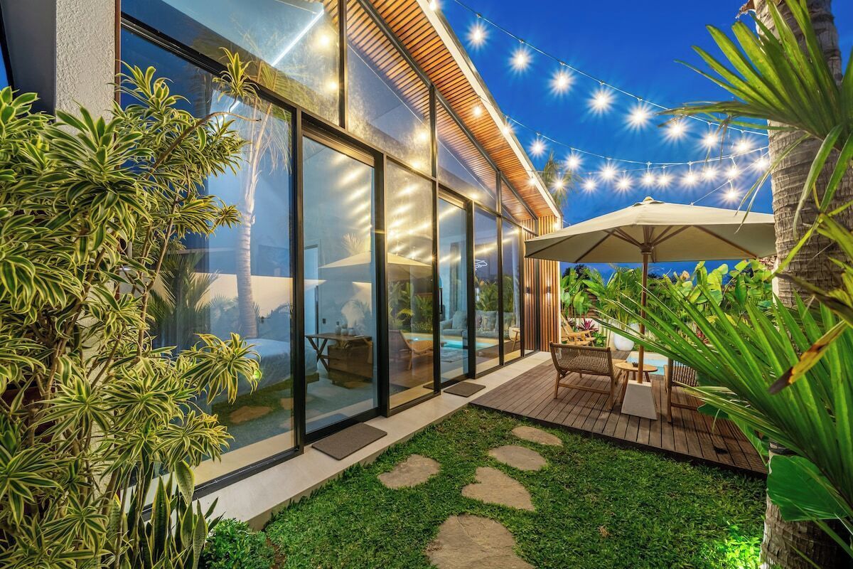 Un espace extérieur moderne doté d’une zone fermée par des parois de verre, d’une terrasse en bois et de guirlandes lumineuses.
